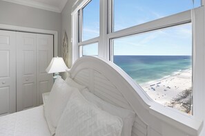 A bedroom with a large window overlooking a beach, a bed with white bedding, and a nightstand with a lamp.