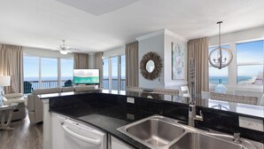 A modern kitchen with a black countertop, stainless steel sink, and a dining area with a view of the ocean. - Sunrise 2011 • Gulf-Front 3BR Retreat • Beach Service (Panama City Beach)