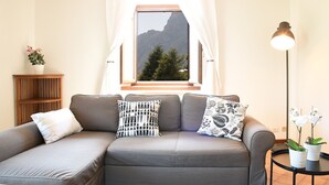 Un sofá gris con cojines estampados, una mesita de madera con una planta en maceta, una lámpara de pie y una ventana con vistas a la montaña.