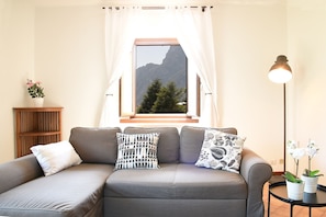 A grey sofa with patterned cushions, a wooden side table with a potted plant, a floor lamp, and a window with a mountain view.