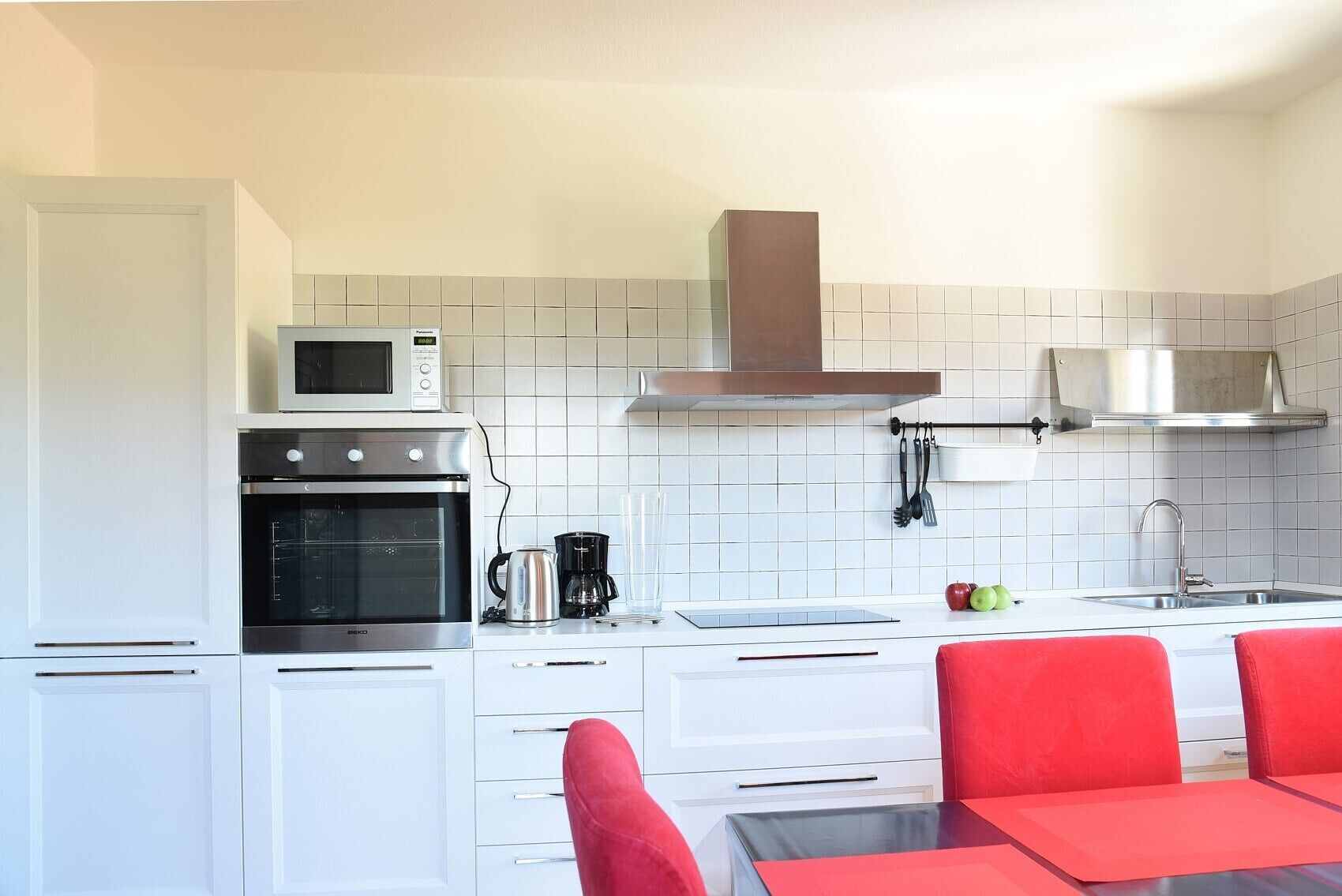 Une cuisine compacte avec des meubles blancs, un four à micro-ondes, un four, une machine à café et un évier. On y trouve des chaises rouges disposées autour d’une petite table.