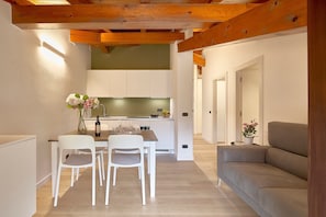Eine moderne Küche mit weißen Schränken, einem Esstisch mit Stühlen und einem grauen Sofa in einem hellen Raum mit Holzbalken.