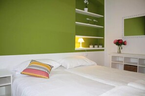 Une chambre à coucher avec un lit, du linge de lit blanc, une table de chevet, une lampe, un miroir et des étagères ornées d’objets décoratifs.