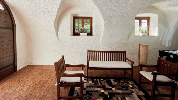 Une pièce avec un mur blanc à la texture particulière, deux fenêtres à cadre en bois, un tapis à motifs et des meubles en bois.