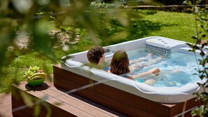 Un bain à remous où se trouvent deux personnes, entouré d’une terrasse en bois et de verdure.