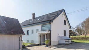 Une maison moderne à deux étages, avec un toit en tuiles, une cheminée bien visible et un petit jardin agrémenté d’une plante en pot.