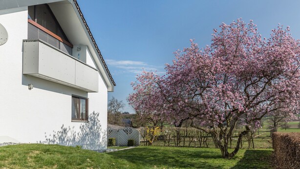 Une maison moderne avec un toit en pente, un balcon et un jardin où les arbres sont en fleurs.