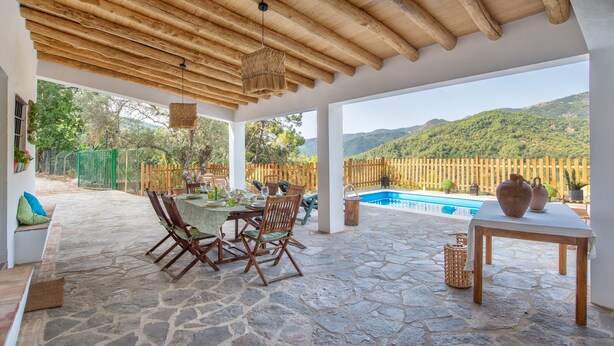 A covered patio with stone flooring, wooden furniture, a pool, and a view of green hills.