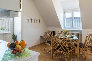 Uma sala de jantar com uma mesa posta para quatro pessoas, rodeada por cadeiras de vime, uma fruteira no balcão e uma janela com persianas.
