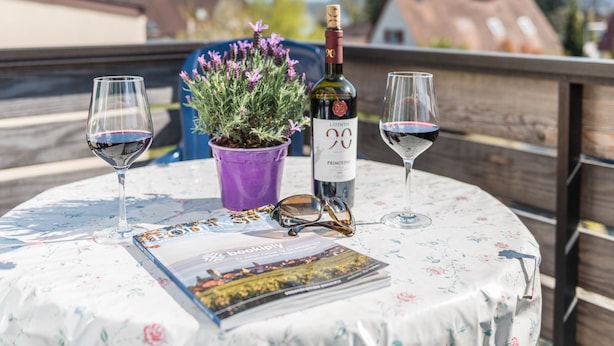 Una mesa redonda con mantel floreado, una botella de vino, dos copas de vino, una maceta morada y una revista.