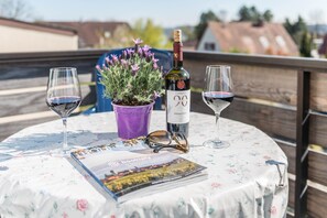 Uma mesa redonda com toalha florida, uma garrafa de vinho, duas taças de vinho, um vaso roxo com flores e uma revista.