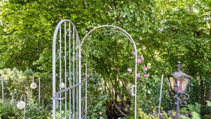 Ein schmiedeeisernes Tor mit bogenförmiger Oberseite überspannt einen Gartenweg, umgeben von üppigem Grün und blühenden Pflanzen.