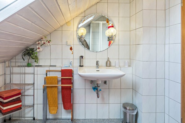 A bathroom with a white sink, a mirror, a towel rack with multiple towels, a trash bin, and a plant.