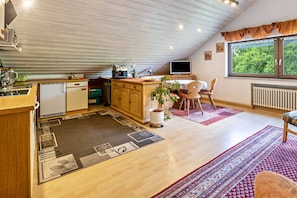 Una cocina compacta con refrigerador, estufa y lavaplatos, una zona de comedor con mesa y sillas, y un ventanal con vistas a la vegetación.