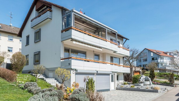 Un edificio residencial moderno de dos plantas con balcón, garaje y jardín ajardinado.