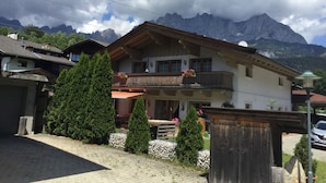 Eine zweistöckige Chalet mit Balkon, umgeben von Grünpflanzen und Bergen im Hintergrund.