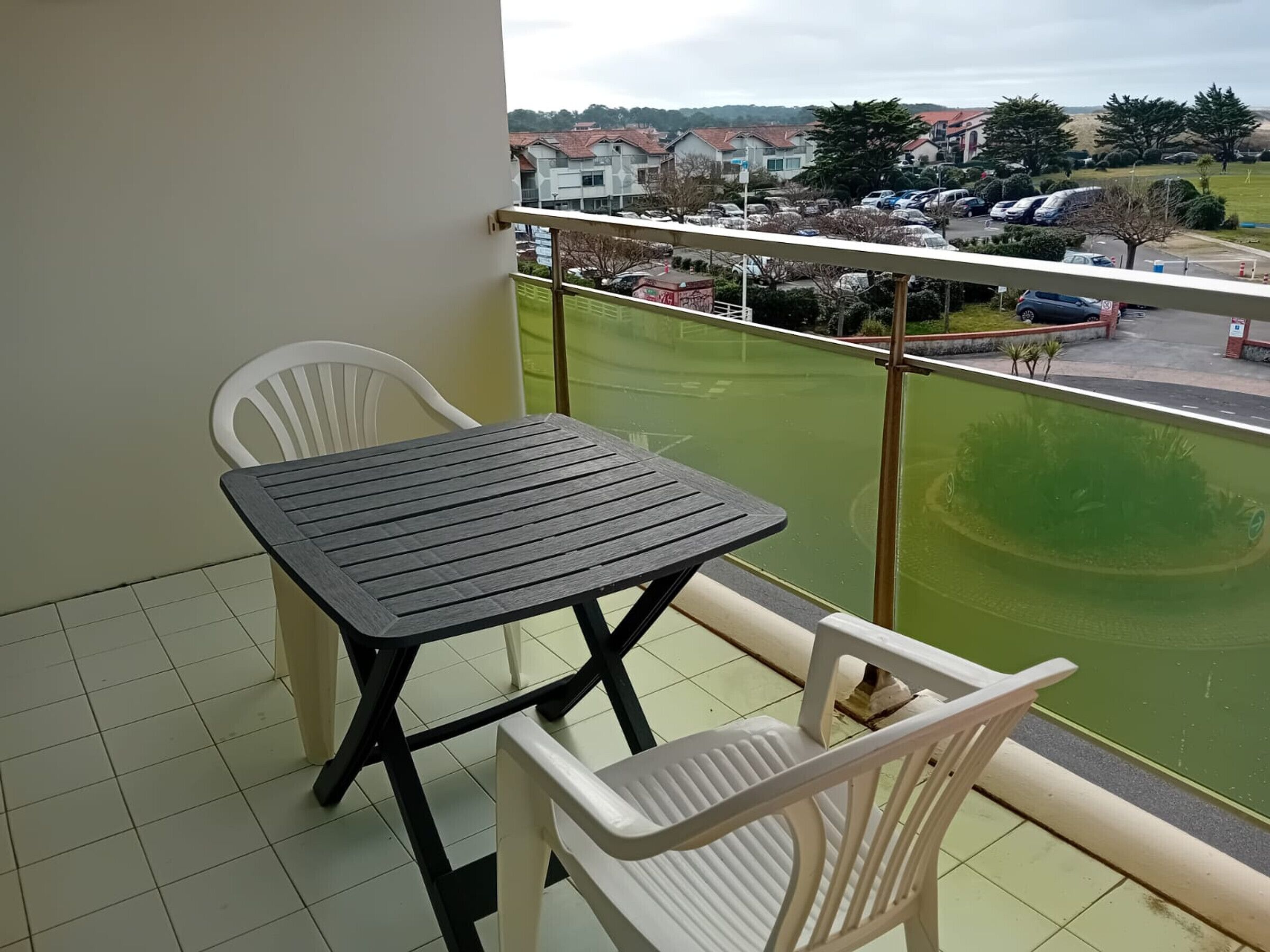 A balcony with a table and two chairs, overlooking a parking area and buildings.