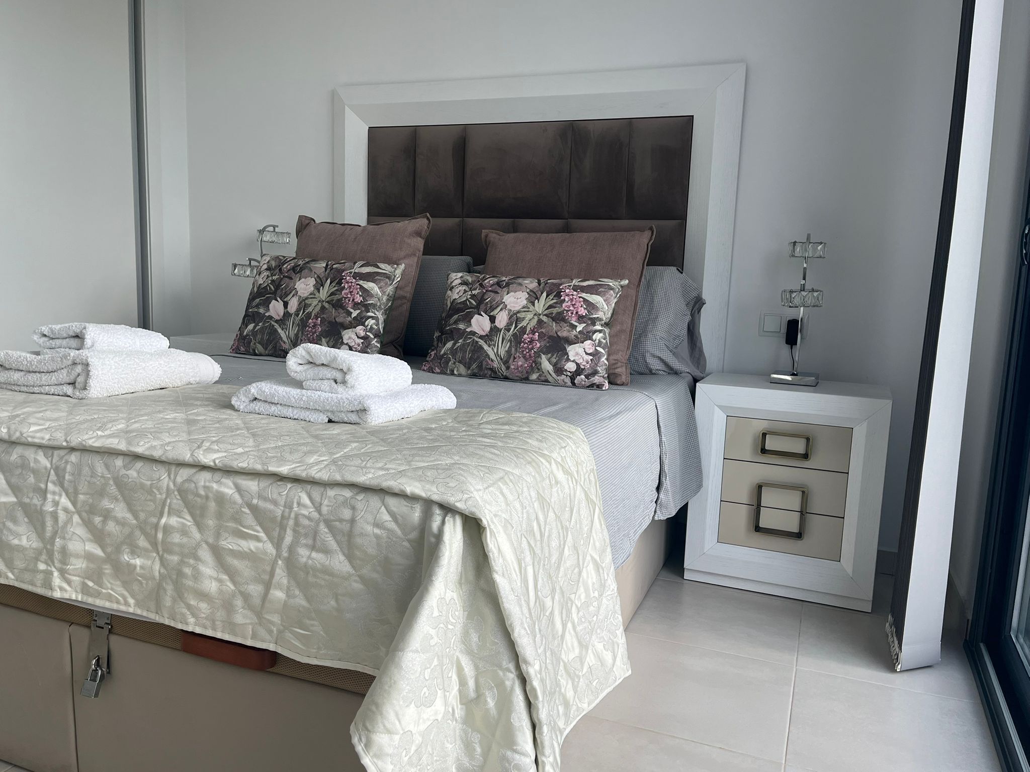 Un lit bien fait, avec une courtepointe, des oreillers et des serviettes pliées. Il y a une table de chevet avec une lampe et un tiroir. La chambre a un design moderne, avec une grande tête de lit et une fenêtre.