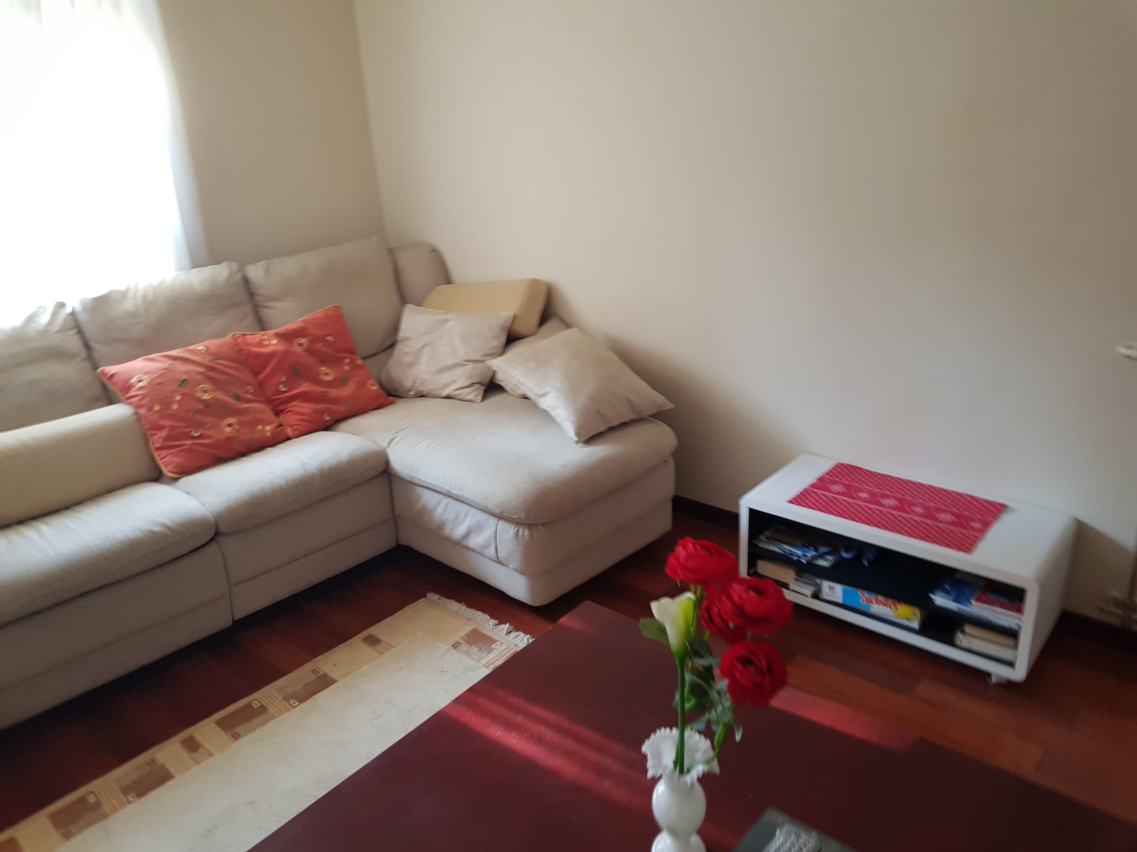 Una sala de estar con un sofá color beige, un cojín rojo, un jarrón blanco con rosas rojas y una mesita auxiliar blanca con revistas.