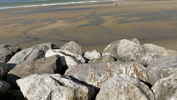 Ein Strand mit großen Felsen im Vordergrund und einem sandigen Ufer, das sich bis zum Meer erstreckt.