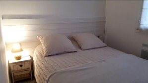 A neatly made bed with white bedding and pillows, a bedside lamp, and a small wooden nightstand with a basket. - T2 Les Lilas ,plage 300m (Hyères)