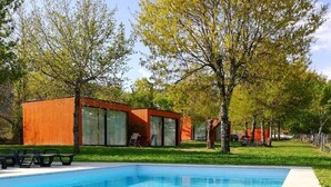 A swimming pool with a clear blue water surface, surrounded by a concrete deck. There are two wooden cabins with large windows and a picnic table nearby.