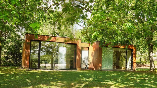 A modern, open-plan house with large glass windows and wooden exterior walls, surrounded by lush greenery.