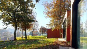A modern building with a glass facade, a red exterior wall, and a grassy area with trees and a fence in the background.