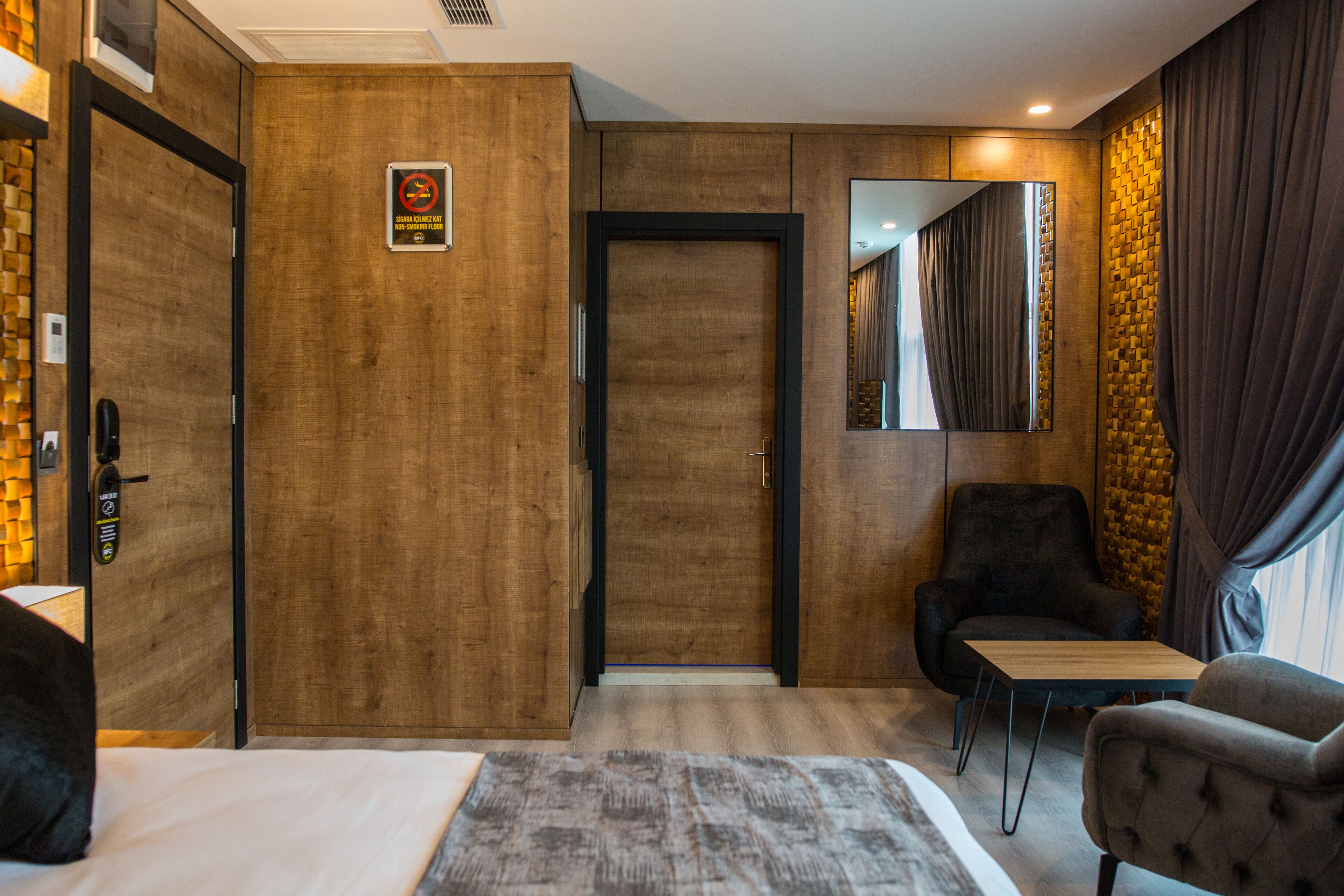 Hotellihuone, jossa on puinen ovi, harmaa matto sängyn alla, pieni pöytä, musta nojatuoli ja peili seinällä.