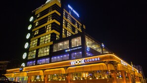 Et hotel med flere etager, hvor et tydeligt 'BFC HOTEL'-skilt er synligt, samt en 'WOOX LOUNGE' i stueetagen og adskillige oplyste vinduer.
