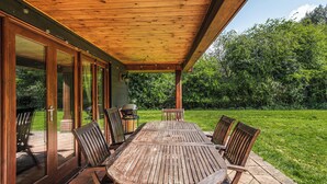 Une terrasse couverte avec une table et des chaises en bois, de grandes portes-fenêtres coulissantes et une vue sur un jardin luxuriant.