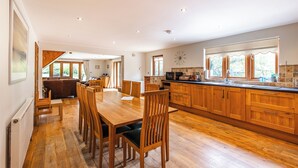Une cuisine spacieuse avec des meubles en bois, un coin repas doté d’une grande table et de chaises, et une vue sur l’extérieur grâce à de grandes fenêtres.