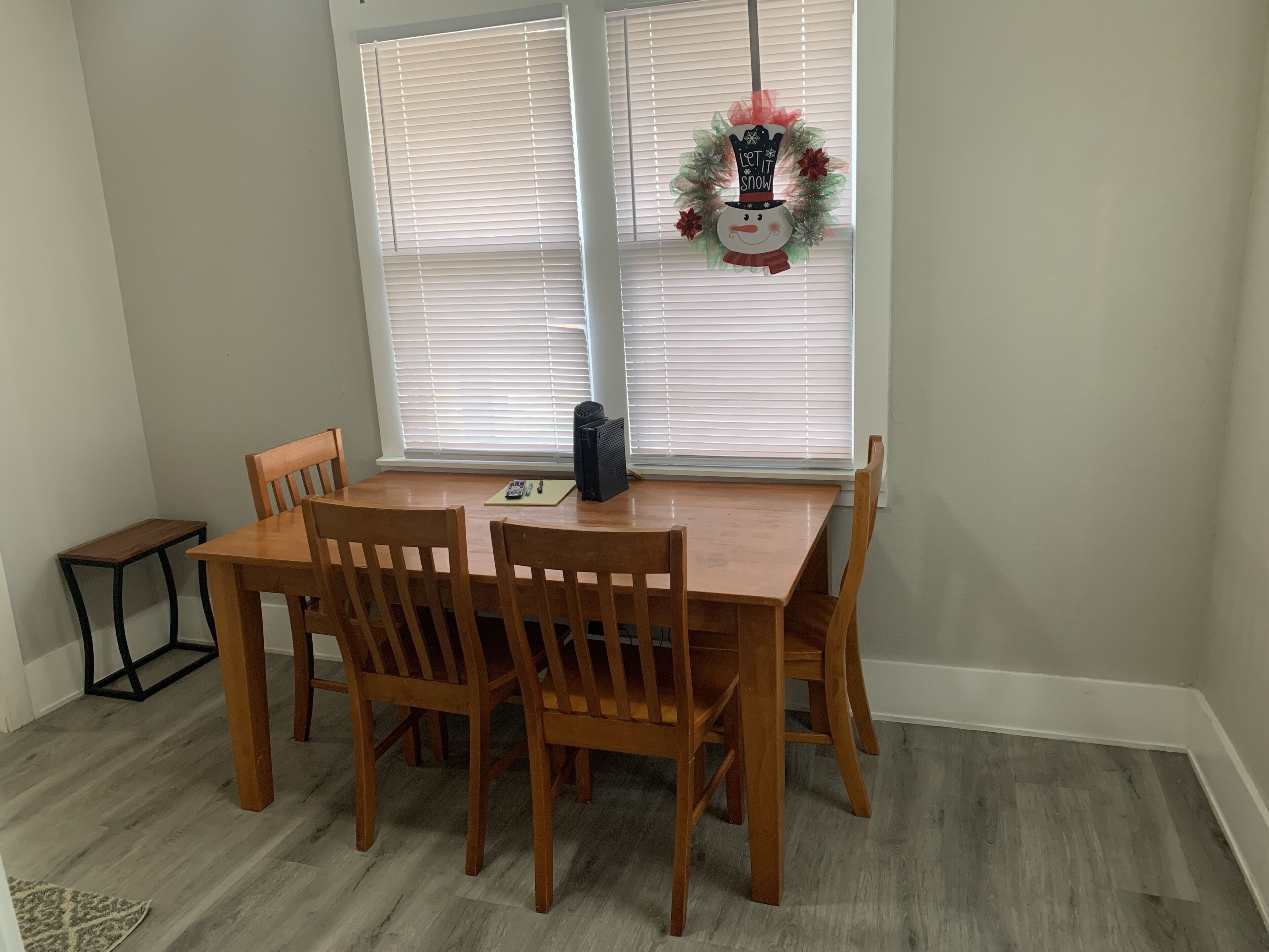 A dining area with a wooden table and chairs, a small side table, and a decorative wreath on the windowsill.