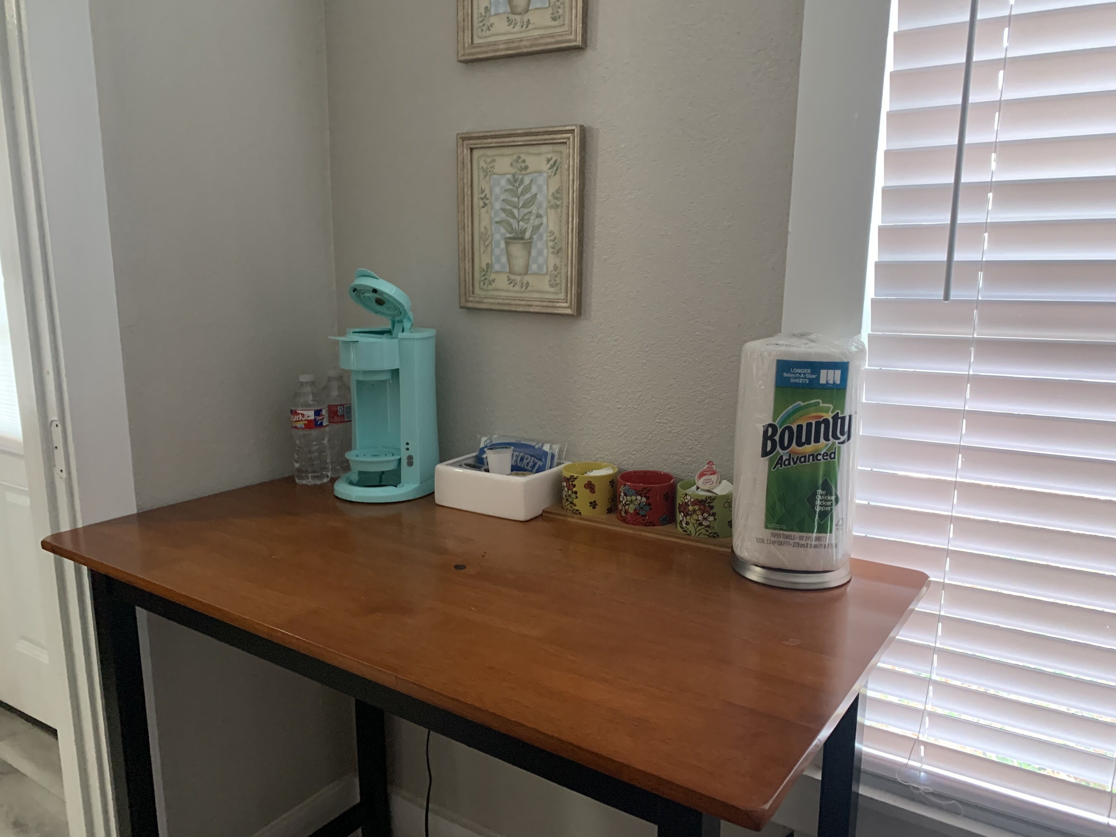 Una mesa de madera con un dispensador de agua, un paquete de toallas de papel Bounty y una hilera de tazas de colores.