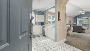 A modern kitchen with white cabinets, a dining area with a gray sofa, and a view through a glass door. - 3br/2ba condo in Barefoot Resort & Golf (North Myrtle Beach, SC)  (North Myrtle Beach)