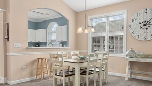 A dining area with a round table, white chairs, a wooden stool, a chandelier, a large mirror, a clock, and a window with blinds. - 3br/2ba condo in Barefoot Resort & Golf (North Myrtle Beach, SC)  (North Myrtle Beach)