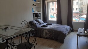 Un dormitorio con cama, una mesa con sillas, una estantería con adornos y una ventana con cortinas.