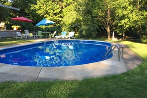 Ein Schwimmbecken mit Betonplattform, umgeben von grünem Gras und Bäumen. Zwei Sonnenschirme und mehrere Liegestühle sind vorhanden.