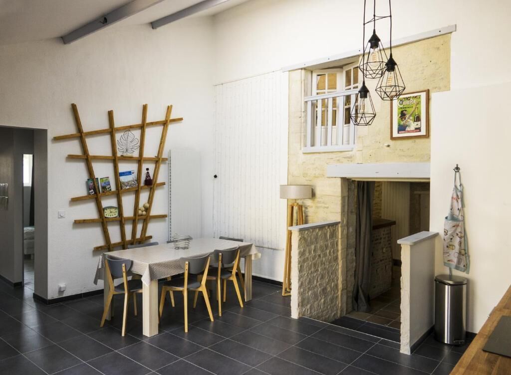 A dining area with a wooden table and chairs, a wall-mounted shelf with books and a plant, and a hanging light fixture.