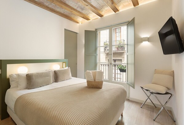 A bedroom with a large bed, a small chair, a TV mounted on the wall, and a window with a view of the outside.