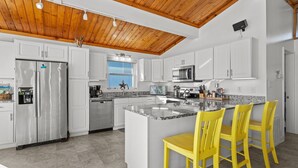 A modern kitchen with white cabinets, a stainless steel refrigerator, a microwave, and a granite island with yellow bar stools. - Pool - Ocean Front - Dogs OK! (Emerald Isle)