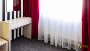 Una habitación con escritorio de madera, un espejo, cortinas rojas y una ventana con cortinas blancas traslúcidas.