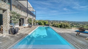 Una terraza de madera con piscina, mobiliario exterior y un edificio de piedra con balcón.