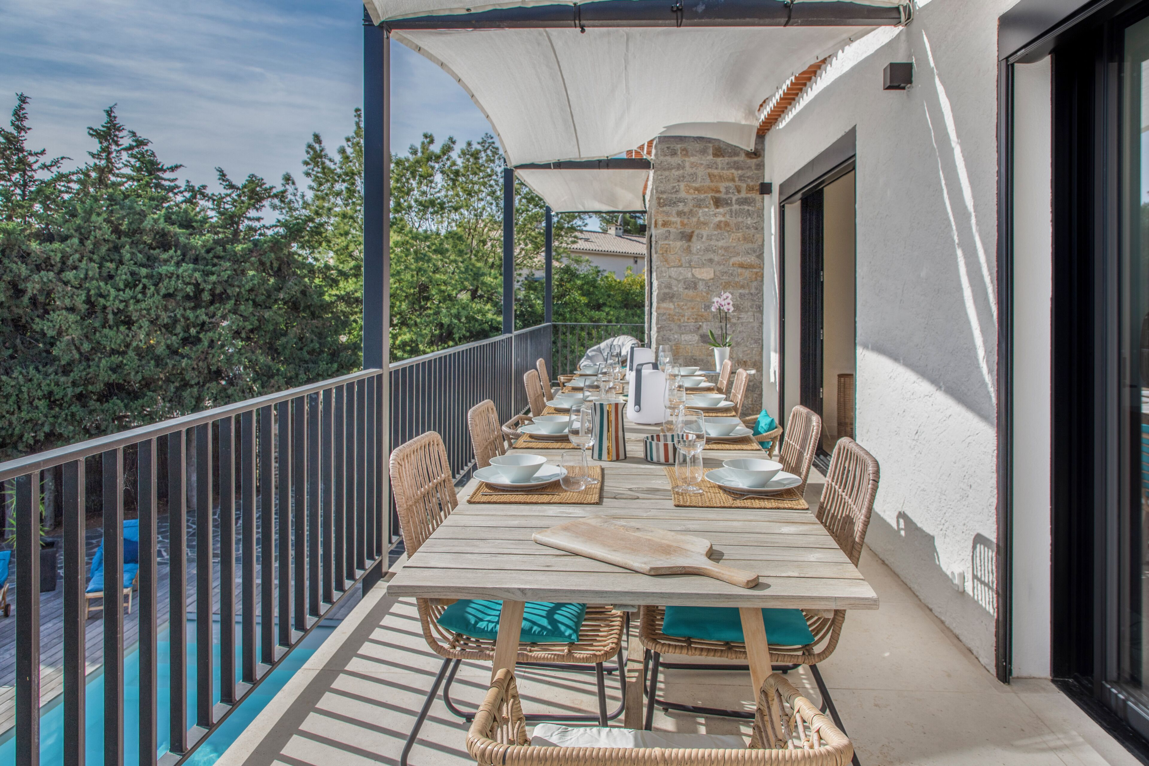 A covered outdoor dining area with wicker chairs, a long table, and a view of trees.