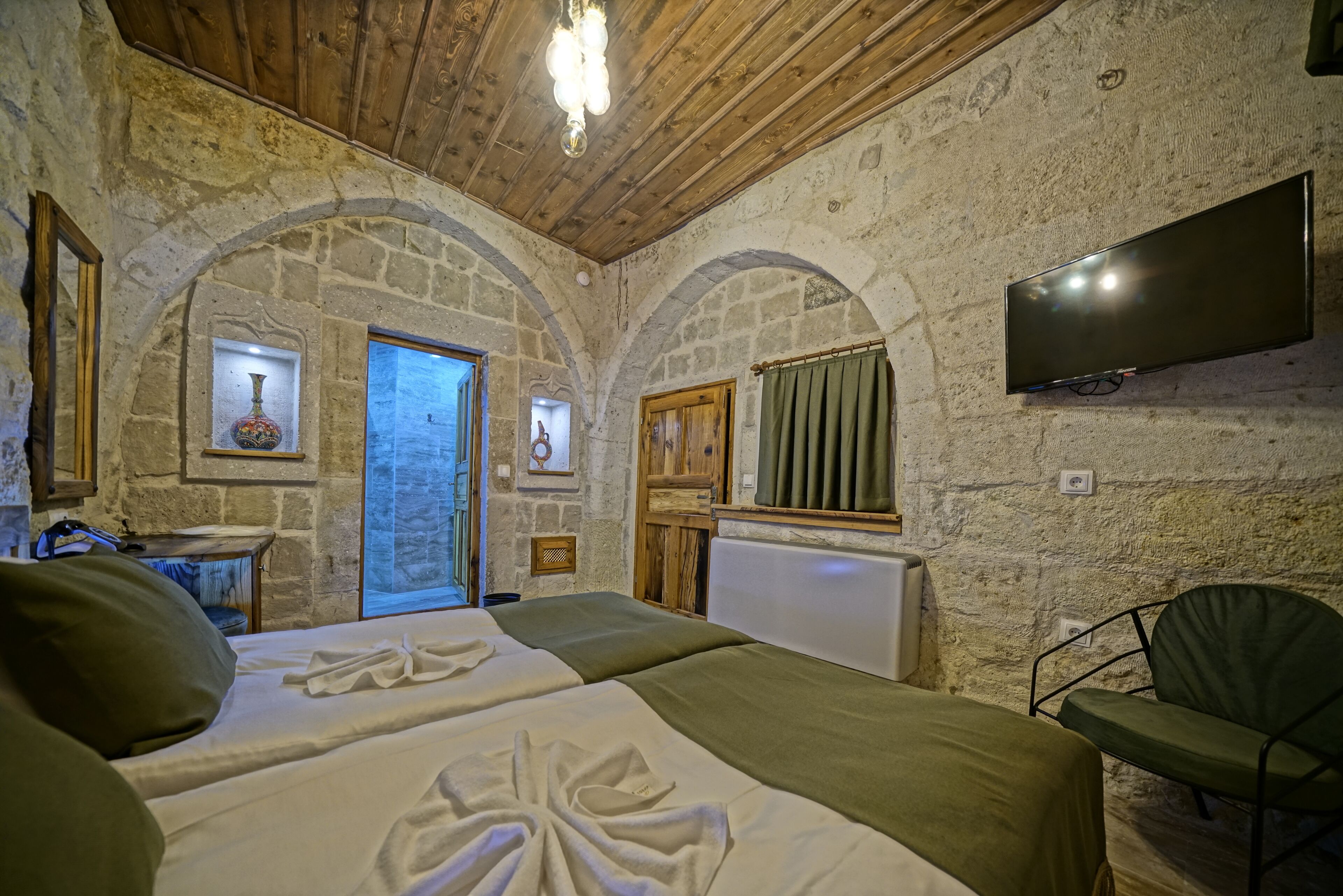 Una habitación con pared de piedra, techo de madera, cama con ropa de cama verde, televisor montado en la pared y una silla.