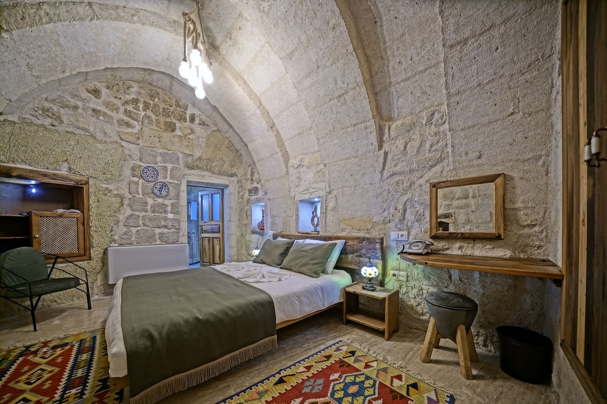 A rustic stone-walled bedroom with a bed, a chair, a nightstand, and a small stool.