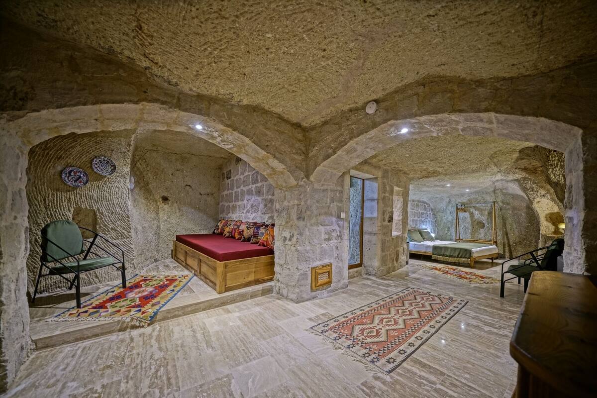 A room with stone walls and vaulted ceilings, featuring two beds with red bedding and a wooden bench.