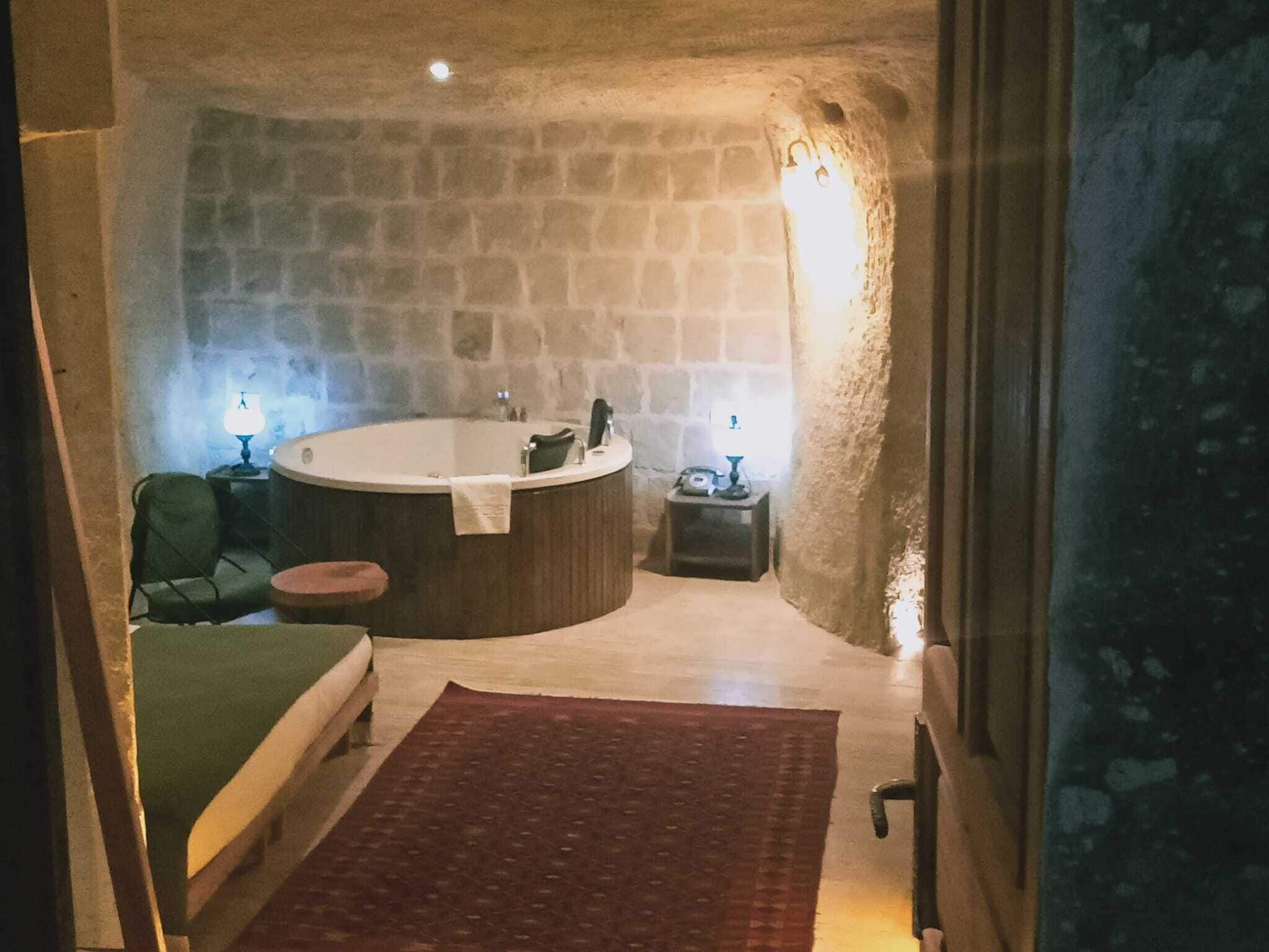 A stone-walled room with a large bathtub, a green chair, a small table with a lamp, and a red rug.