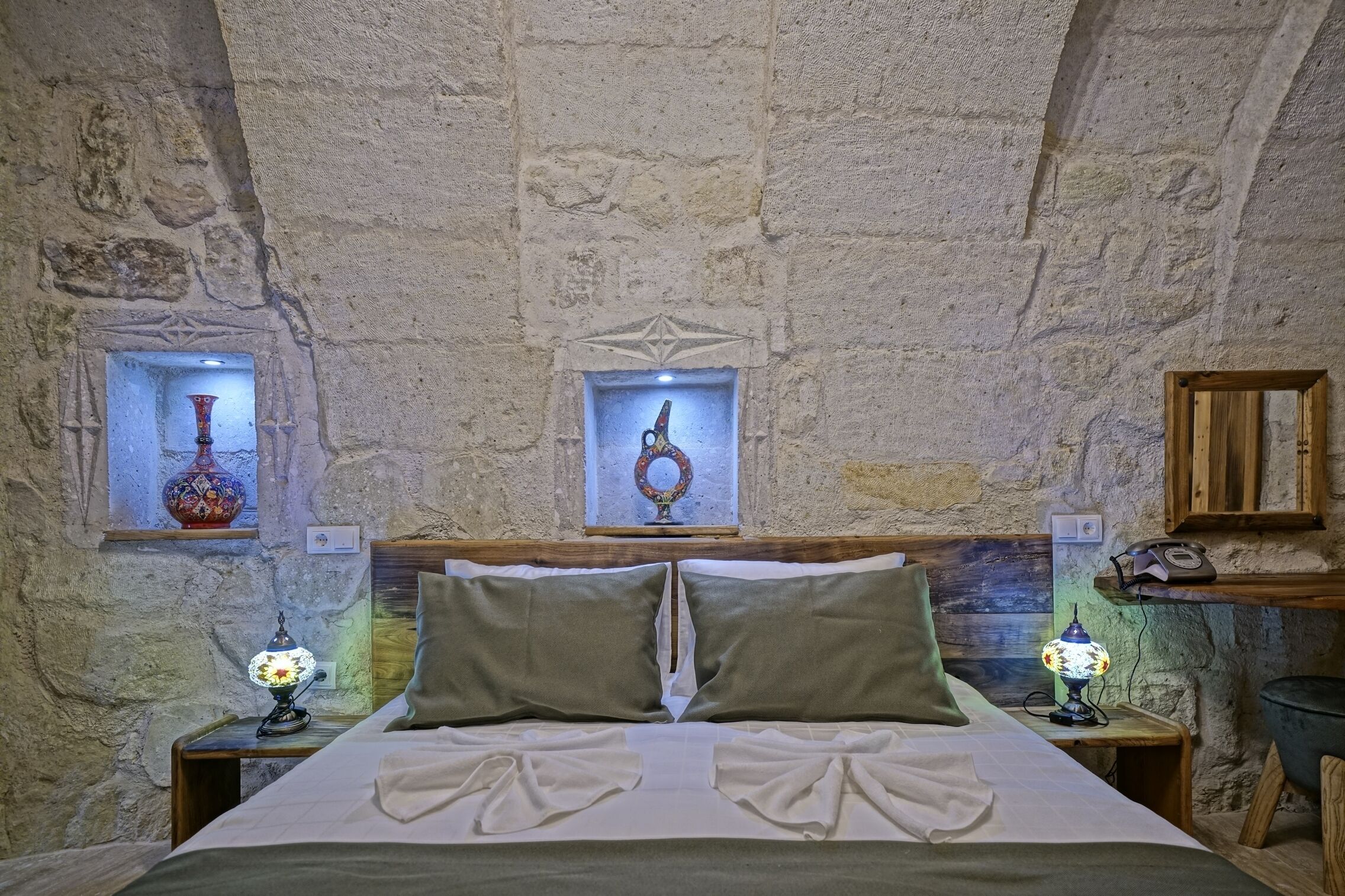 A stone-walled room with a bed, two nightstands, and two wall-mounted windows with decorative items.