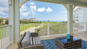 A balcony with wicker furniture, a glass table, and a view of a golf course and ocean.